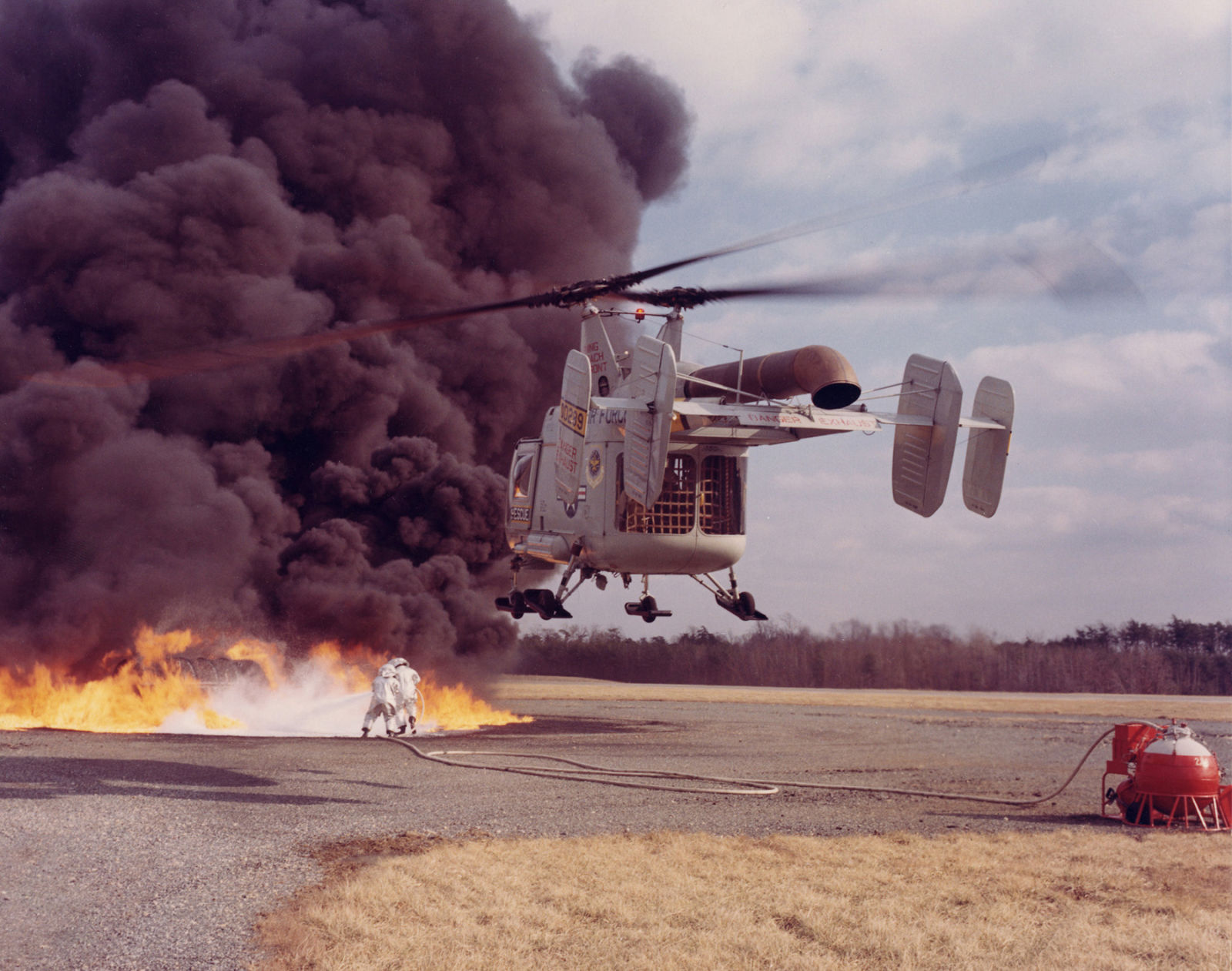 An HH-43B participates in an firefighting exercise. The crew of the helicopter uses the rotor downwash to open a path for rescuers to spray foam. 