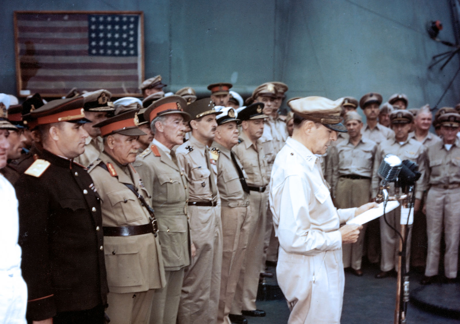 The Perry flag is visible in the background behind the assembled military dignitaries. General MacArthur is at the microphone. 